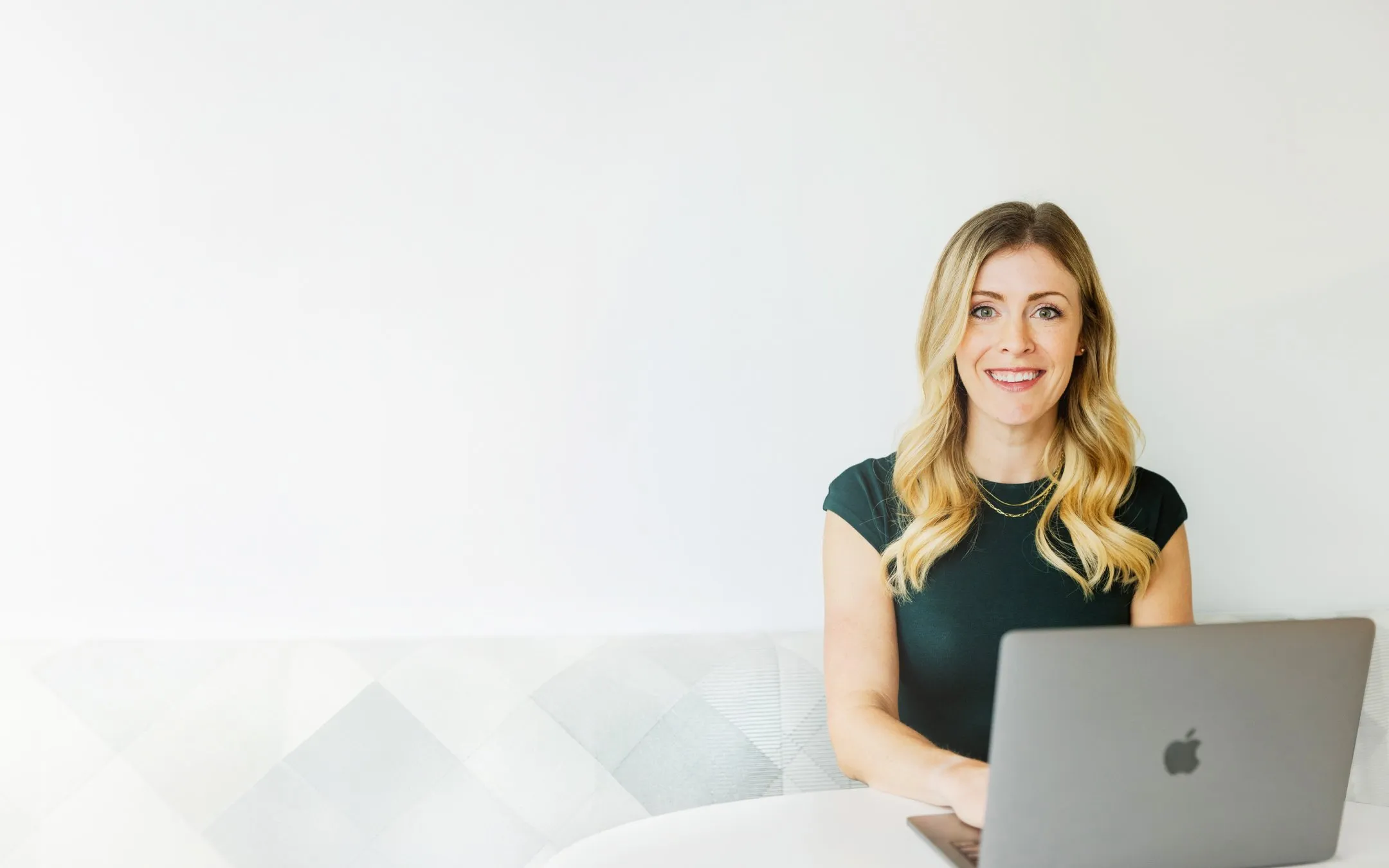 woman smiling in front of laptop sitting on a gray couch with a blank wall behind her